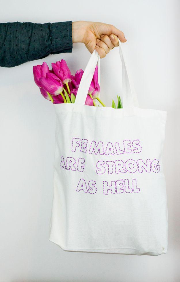 White canvas tote held by one hand, stitched with purple outlined block capitals, pink tulips in the top of the bag