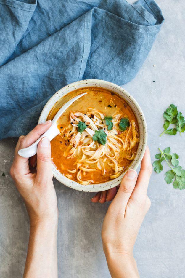 Hands holding a speckled bowl of vegan Thai coconut soup with rice noodles and cilantro on a grey surface