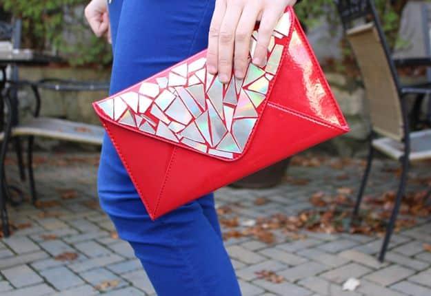 Red patent envelope clutch with silver CD shards glued across the flap, held by a woman in blue jeans on a patio