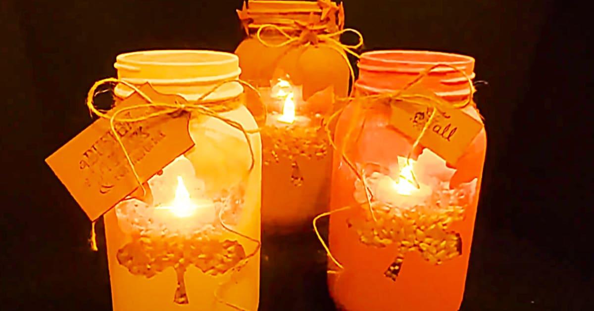 Row of orange mason jar candle holders with paper leaf tags and lace, lit from inside against a dark wall
