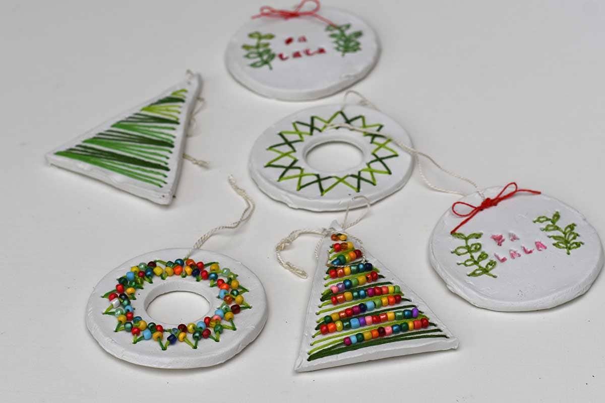 Six white air dry clay ornaments in tree, wreath and round shapes stitched with green thread, red letters and colorful beads
