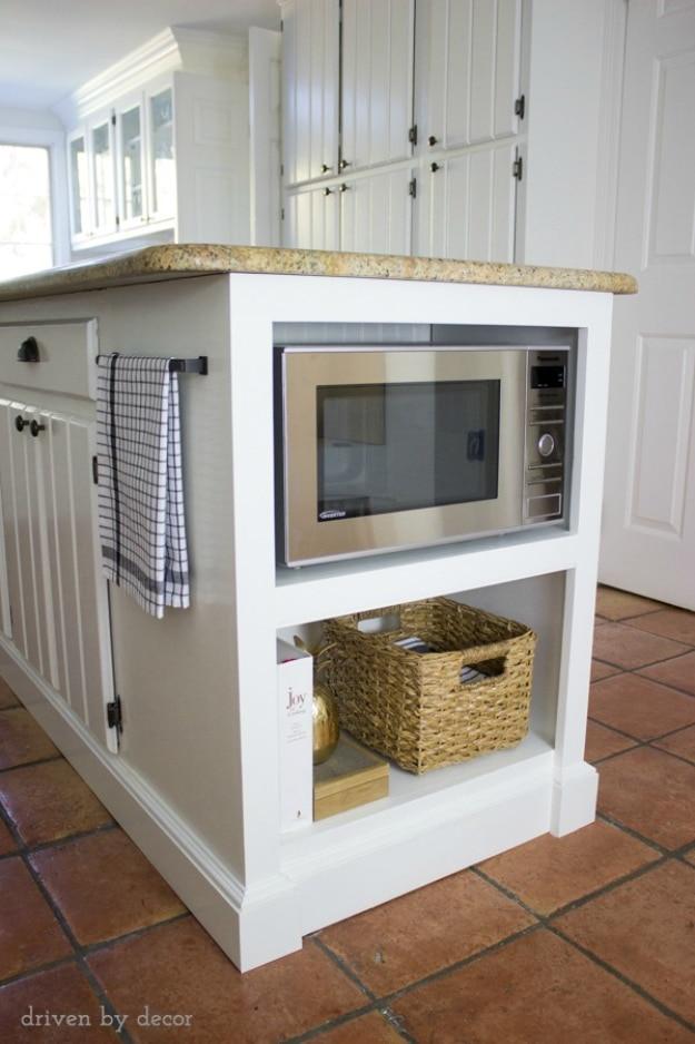 White kitchen island end with a stainless microwave built into a framed recess above an open shelf holding a woven basket