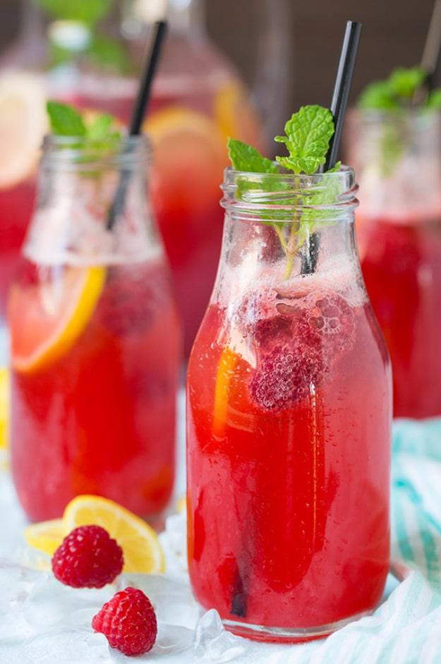 Two glass bottles of ruby pink raspberry lemonade with black straws, mint sprigs, lemon slices and berries