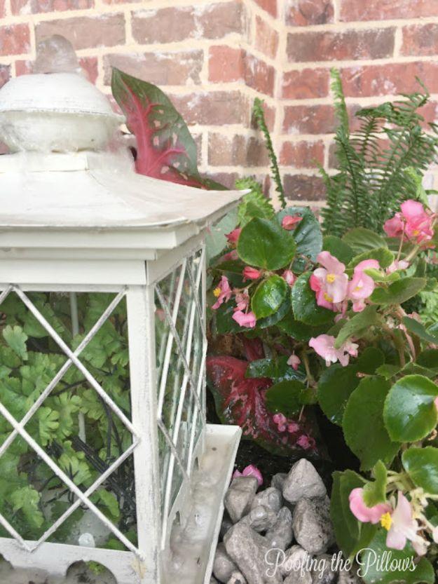 White lantern style bird feeder fountain set among pink begonias and stones against a red brick wall