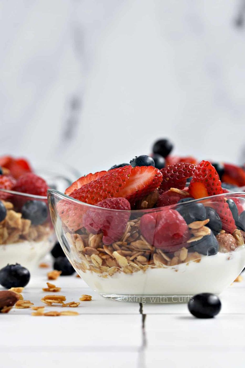 Footed glass dish of yogurt parfait with granola over white yogurt and berries heaped on top, a second dish blurred behind