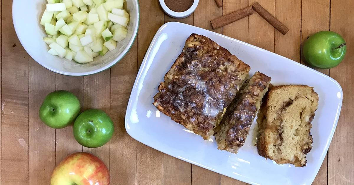 Apple Bread - sliced loaf with a cinnamon streusel top and white glaze on a white platter, ringed by whole and diced green apples and cinnamon sticks
