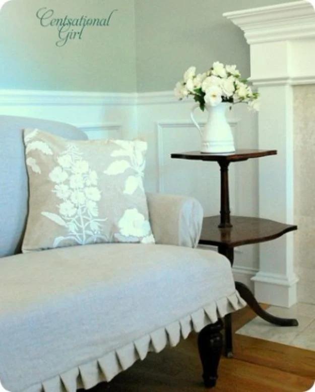 Small settee slipcovered in pale linen beside a dark wood table, styled with a white embroidered floral pillow