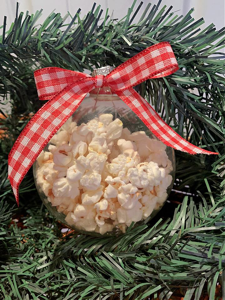 A clear glass ball ornament filled with popped popcorn and topped with a red gingham bow hanging in artificial greenery