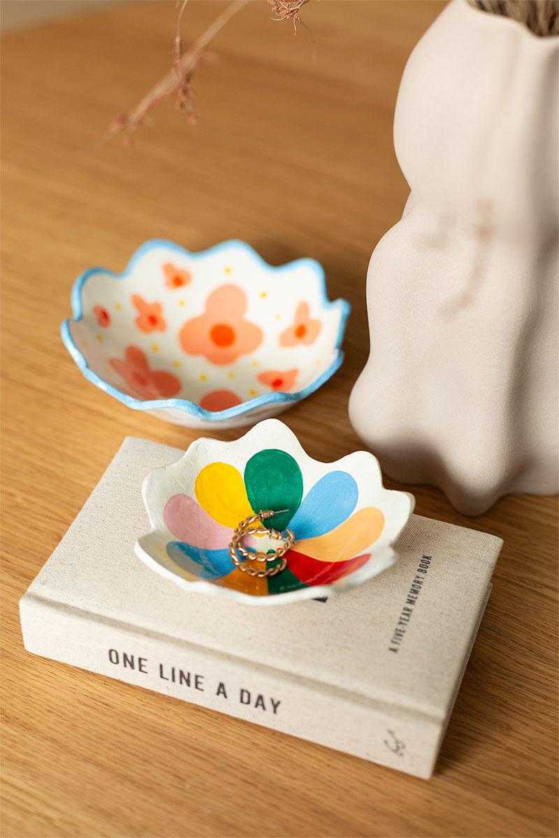 Two wavy edged white clay trinket dishes painted with bright flowers, one holding a gold hoop earring