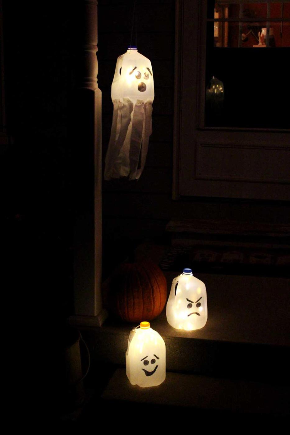 Two sitting recycled milk-jug ghost luminaries glowing with battery lights beside a pumpkin on a dark porch
