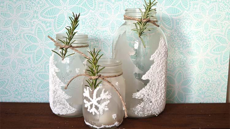 Three frosted mason jars with white Epsom salt pine tree and snowflake designs, tied with twine and rosemary sprigs
