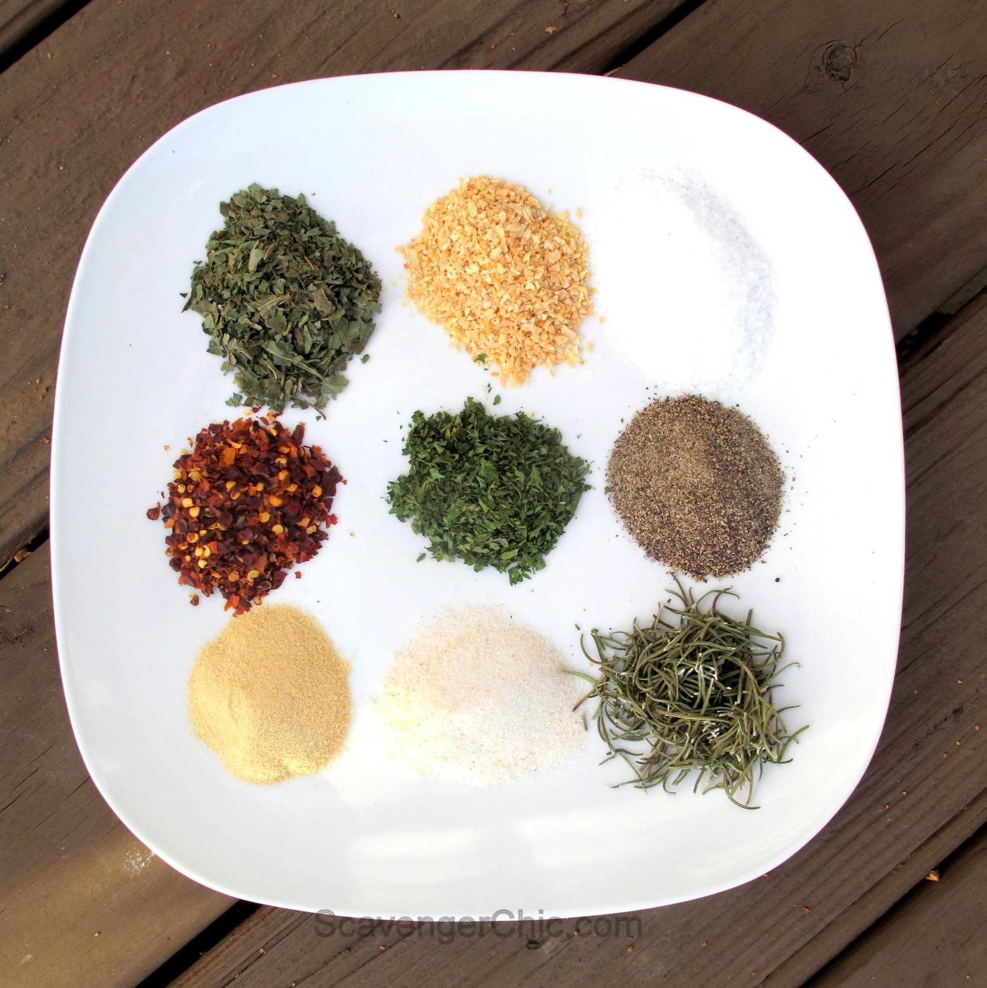 Nine small piles of dried basil, parsley, rosemary, red pepper, garlic, salt and pepper on a white plate on a wood deck