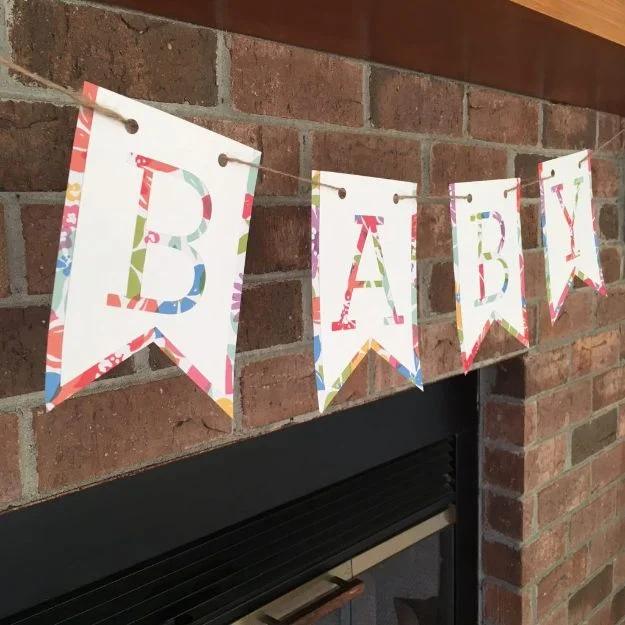 DIY baby shower decoration banner spelling baby in floral print letters on white pennant flags across a brick fireplace
