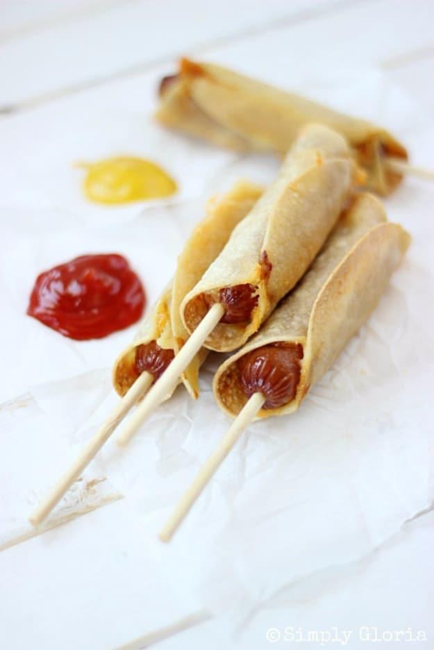 Three corn tortilla wrapped cheese dogs on short wooden skewers with mustard and ketchup on white paper