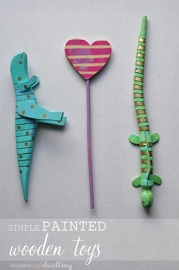A teal painted wooden alligator with gold dots, a striped green lizard and a pink striped heart on a stick on gray