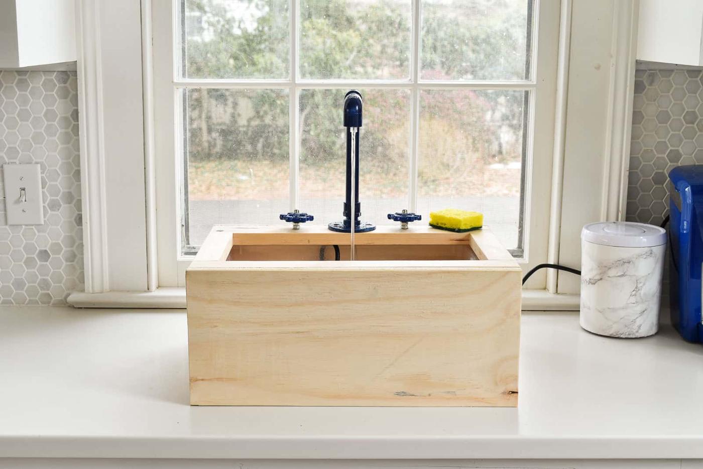 A plain wood toy sink with a blue pipe faucet running water and two blue knobs sitting on a counter in front of a window