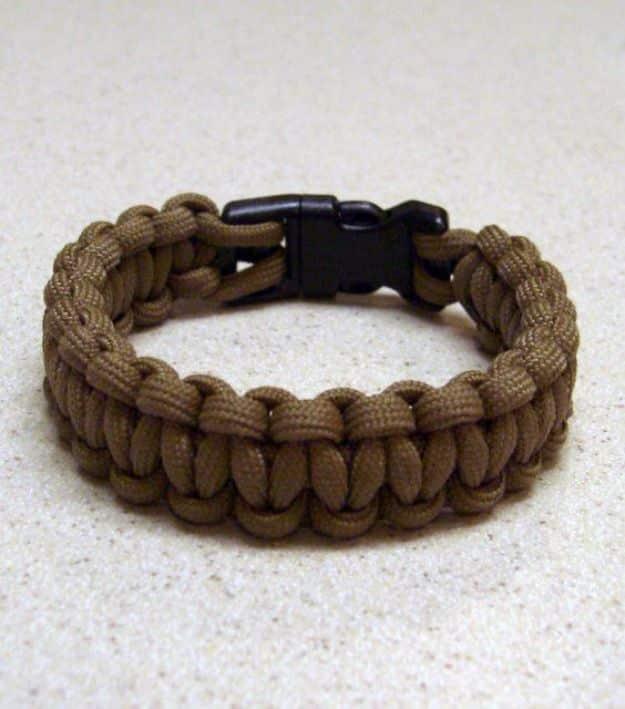 Brown cobra weave survival bracelet with a black plastic side release buckle lying on pale sand grains