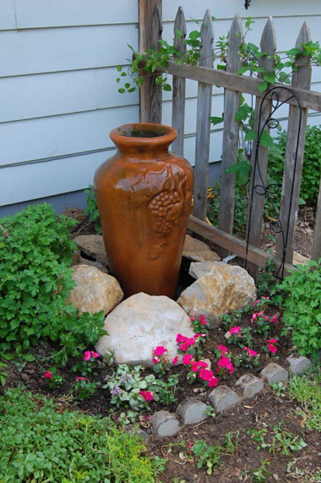 Tall amber glazed urn trickling quietly beside a fence over pale rocks and a bed of bright pink flowers