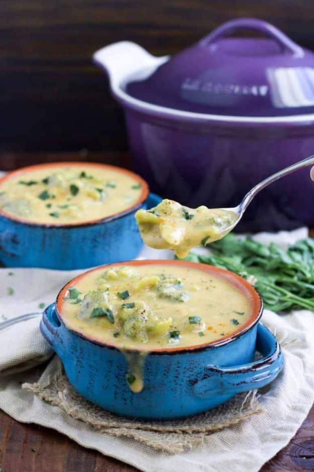 Two blue crocks of vegan broccoli cheese soup with a spoonful lifted, a purple dutch oven in the background