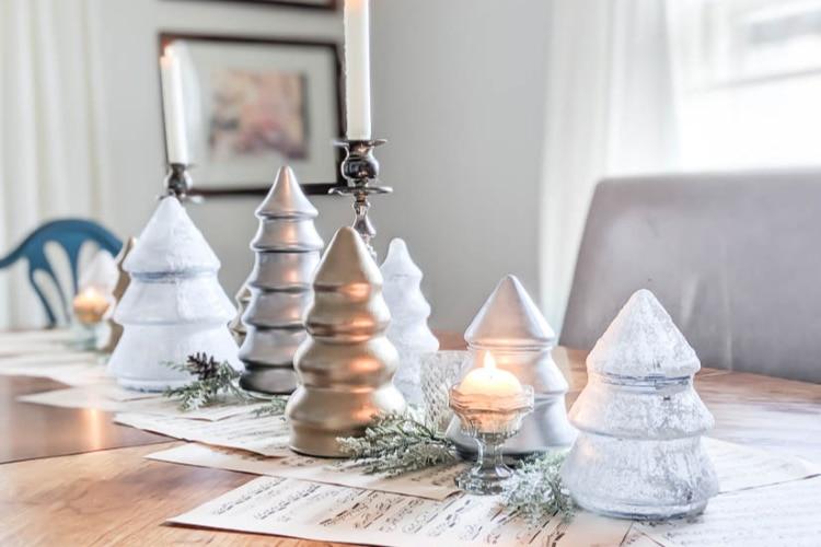 A row of thrifted glass Christmas tree jars painted gold, silver and frosted white with a candle on a sheet music runner