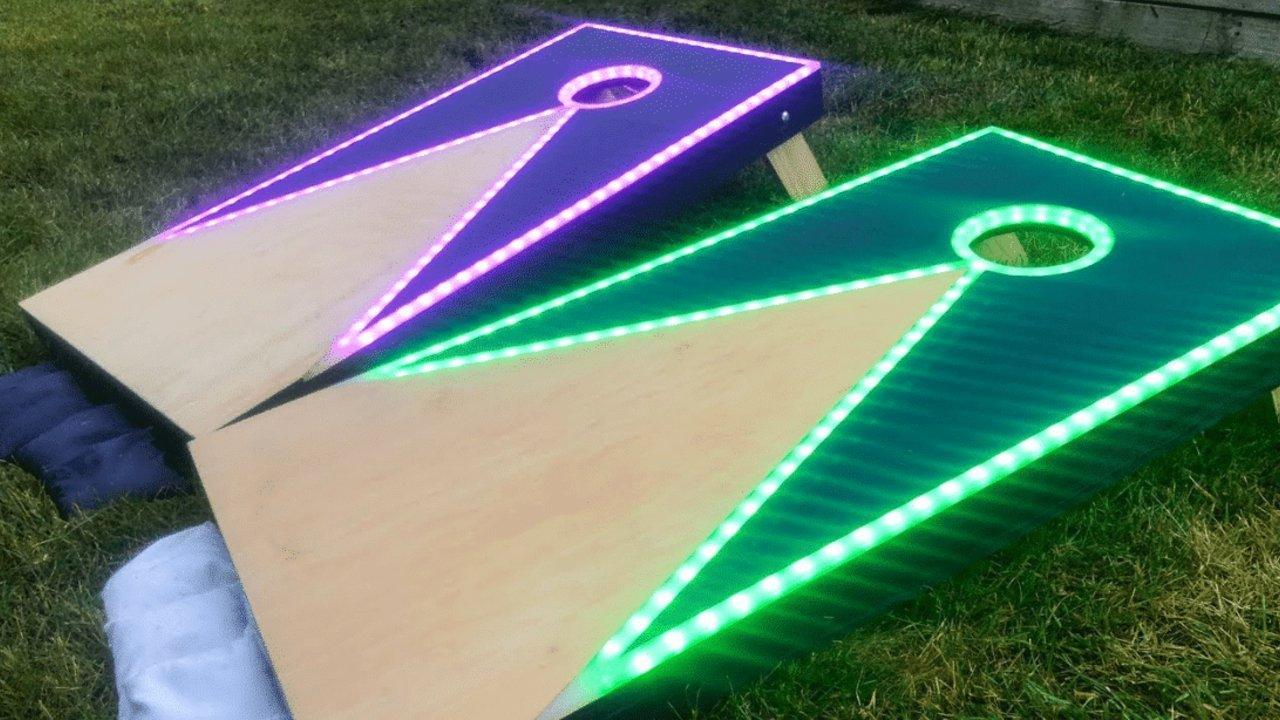 Two DIY cornhole boards on grass outlined in purple and green LED strips glowing around the hole and the edges