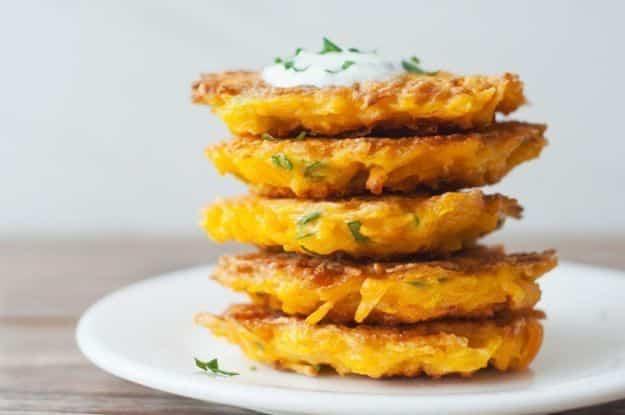 Butternut squash fritters stacked five high on a white plate topped with sour cream and chopped herbs