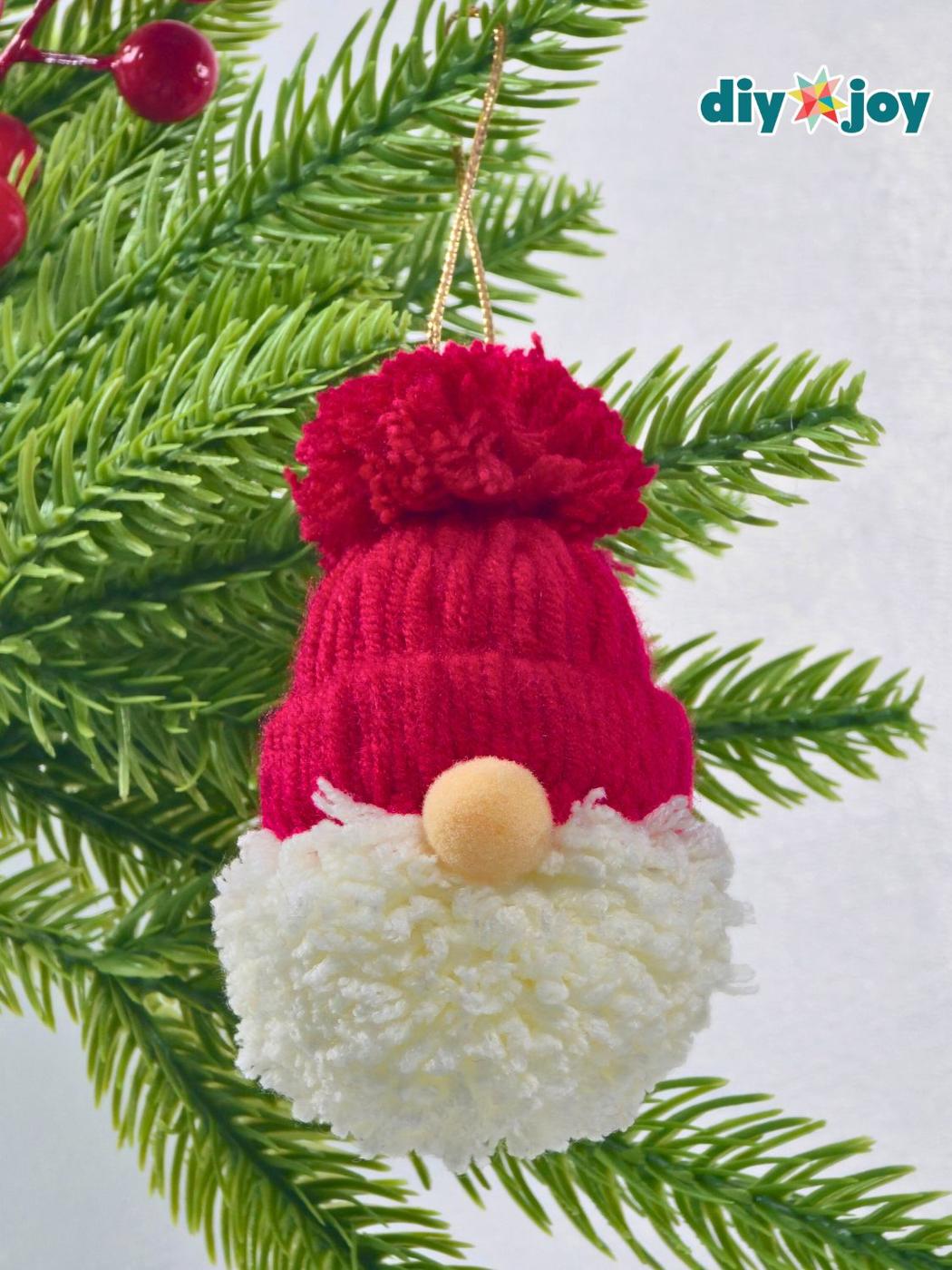 A Santa gnome ornament with a red yarn hat and pom pom, a white yarn beard and a peach pom pom nose hanging on gold cord