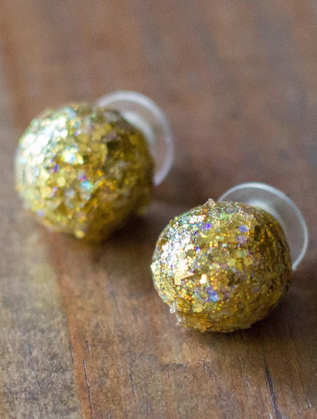 Gold glitter ball stud earrings, two round chunky gold glitter balls on clear plastic posts, on a dark wood table