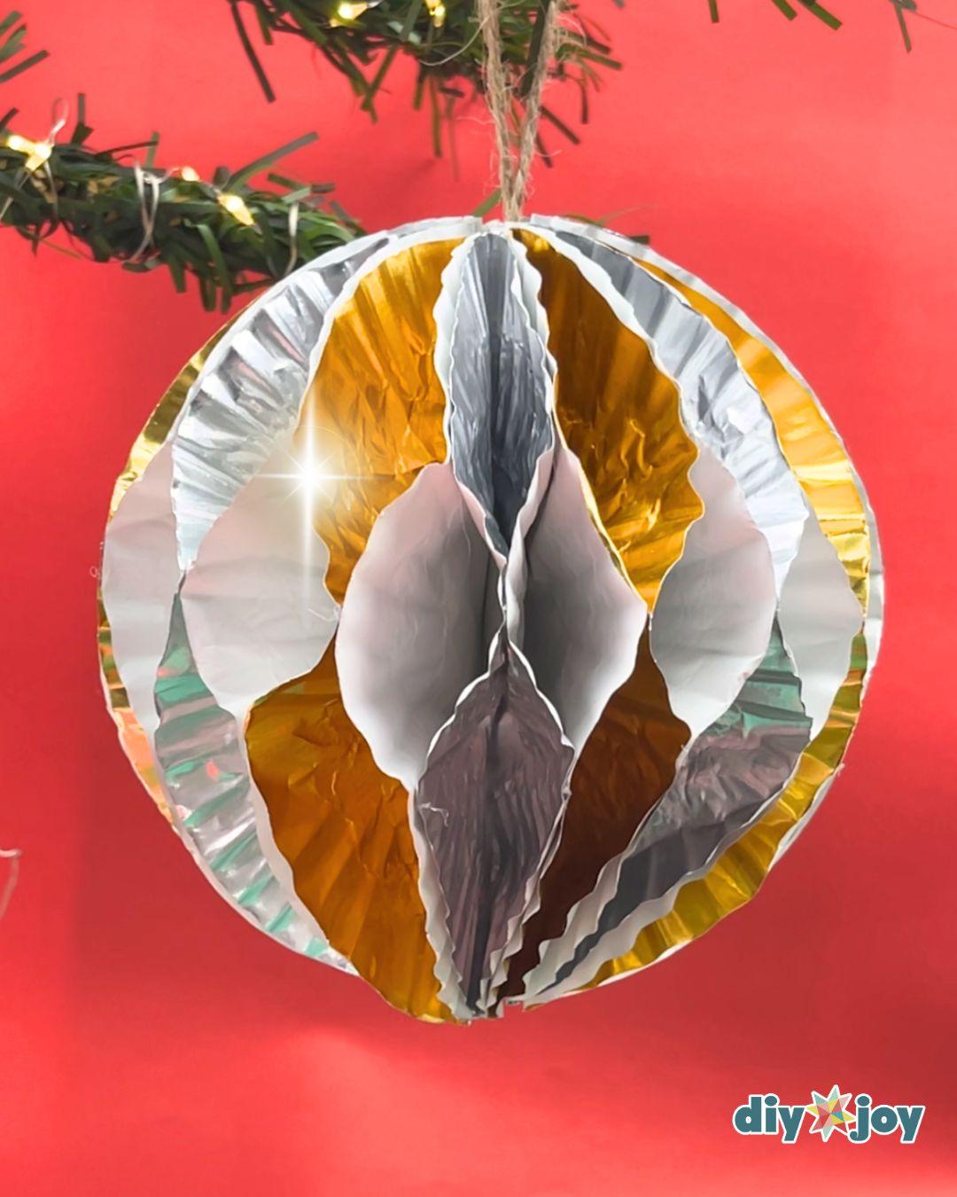 Homemade Ornaments - a finished gold, white, and silver striped cupcake liner ornament hanging in front of a red backdrop and lit garland