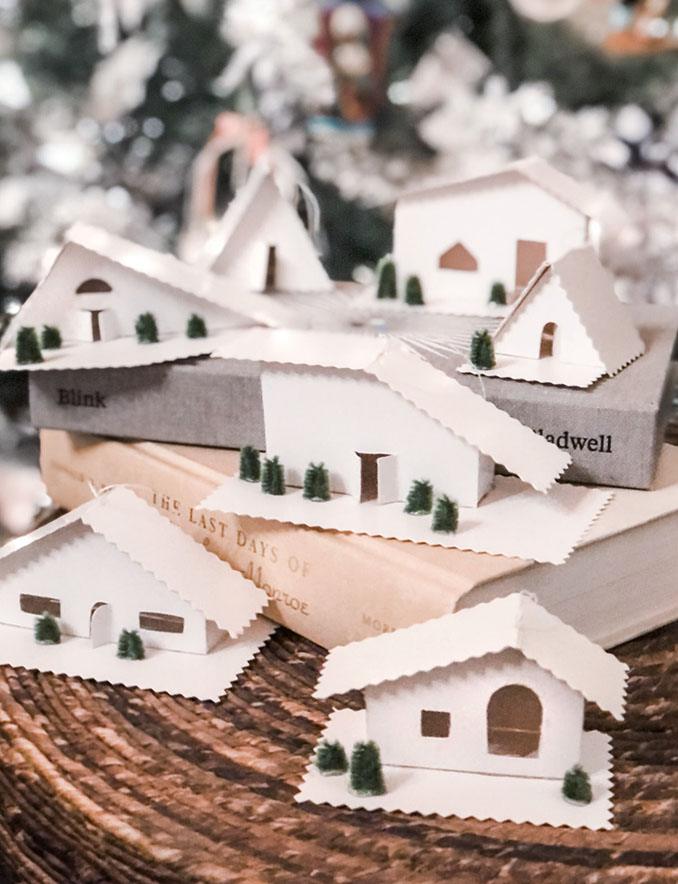 Seven small white cardboard house ornaments with tiny green trees and scalloped roofs displayed on a stack of books