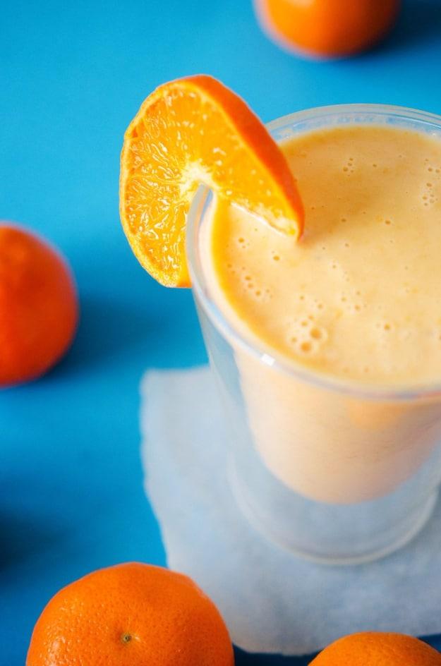 Orange creamsicle smoothie in a glass with an orange wheel on the rim against a bright blue table scattered with oranges