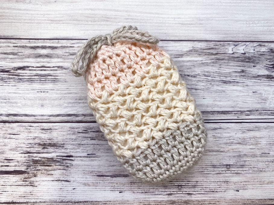 Crochet soap saver bag striped in peach, cream and gray cotton with a drawstring top on a white wood surface