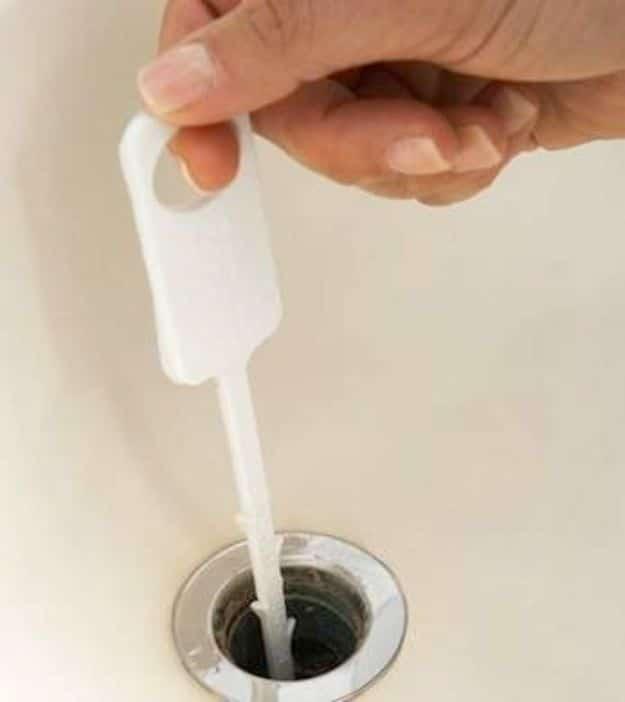 Hand holding a white plastic barbed drain cleaning strip being fed down into an open bathroom sink drain