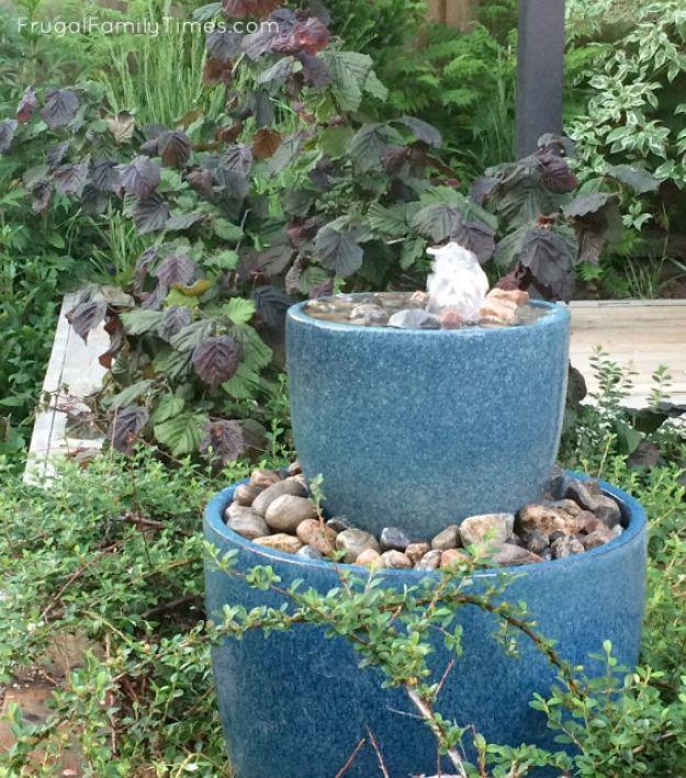 Two blue glazed pots stacked into a bubble fountain ringed by pebbles and leafy green garden planting