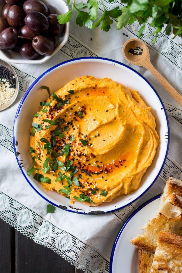 Bowl of orange pumpkin hummus swirled and topped with herbs and sesame seeds beside olives and flatbread
