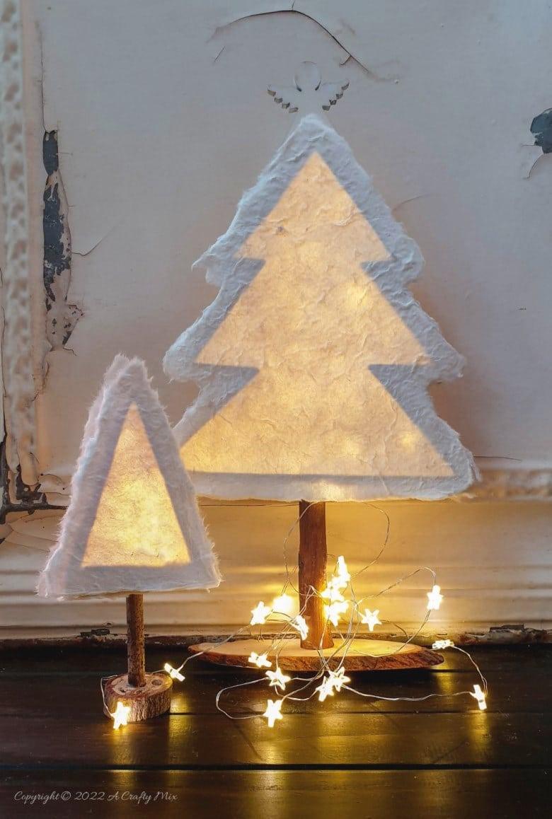Two white handmade paper Christmas trees on twig trunks glowing from fairy lights inside, standing on wood slices