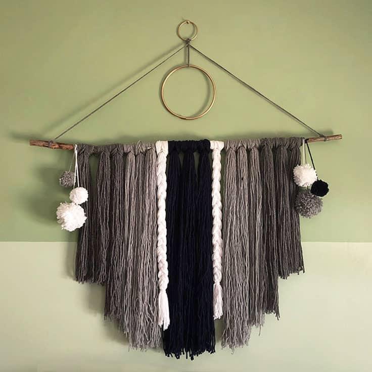 Bedroom wall art idea, a boho yarn wall hanging with gray and black fringe, braids and pom poms on a branch