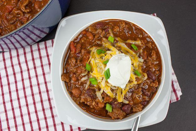 Thick homestyle beef chili in a white bowl with sour cream and green onion on a red checked tablecloth