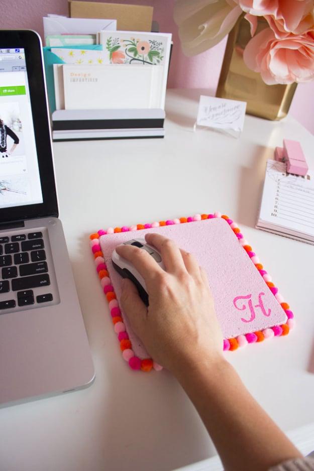 Pom pom mouse pad, a pink square cork mouse pad with a monogram and pink pom pom trim under a hand on a computer mouse