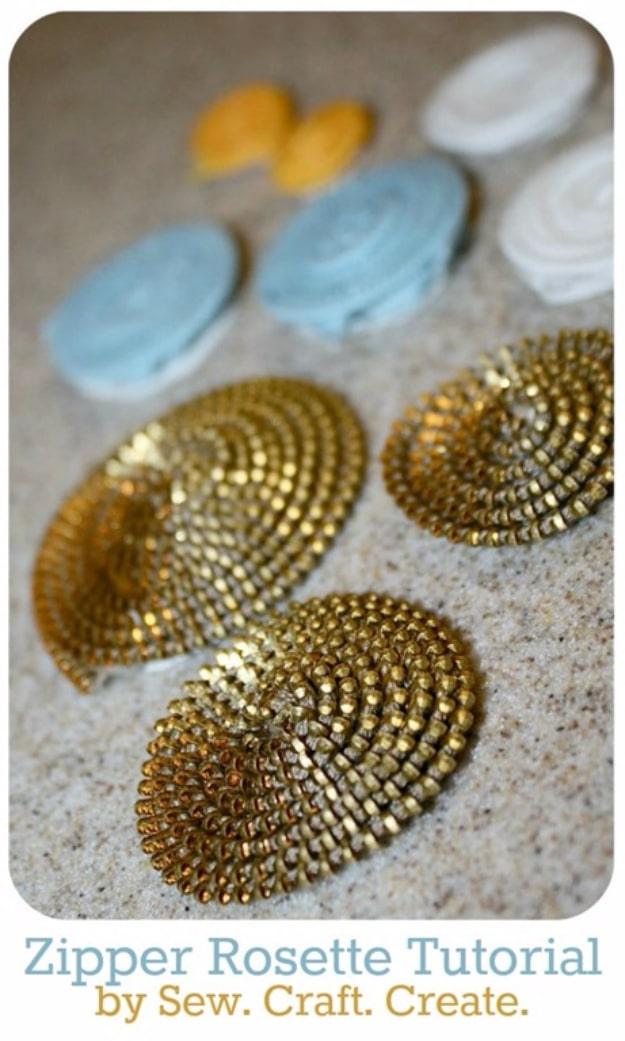 Three gold zipper rosettes with spiral rows of metal teeth resting on sand beside pale blue and white fabric rosettes
