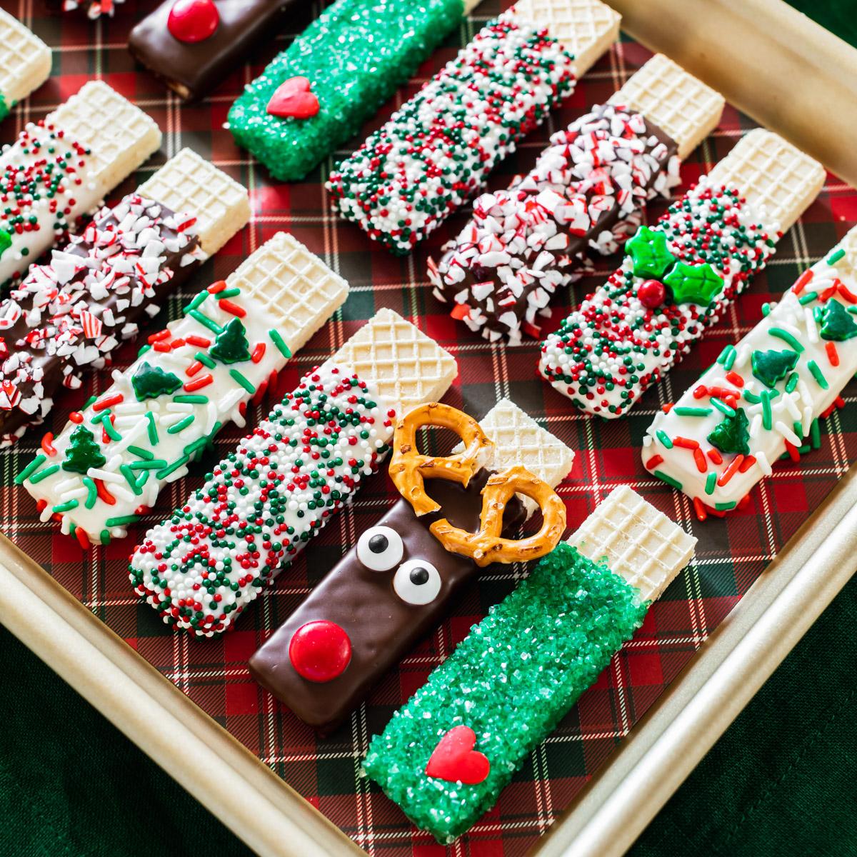 A tray of chocolate dipped sugar wafers decorated with sprinkles, crushed peppermint, green sanding sugar and a reindeer face