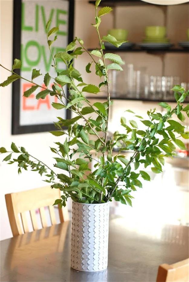 Penny craft vase sprayed white over rows of glued coins, holding green branches on a sunlit dining table