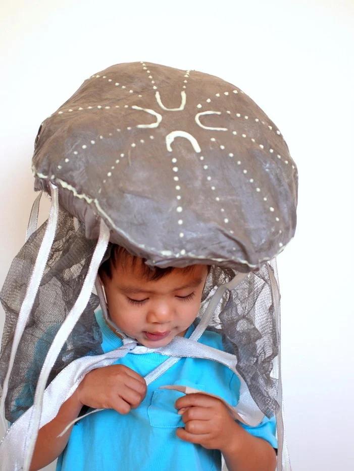 Child wearing a glow-in-the-dark jellyfish hat with a bubble-wrap dome, painted details, mesh and silver ribbons