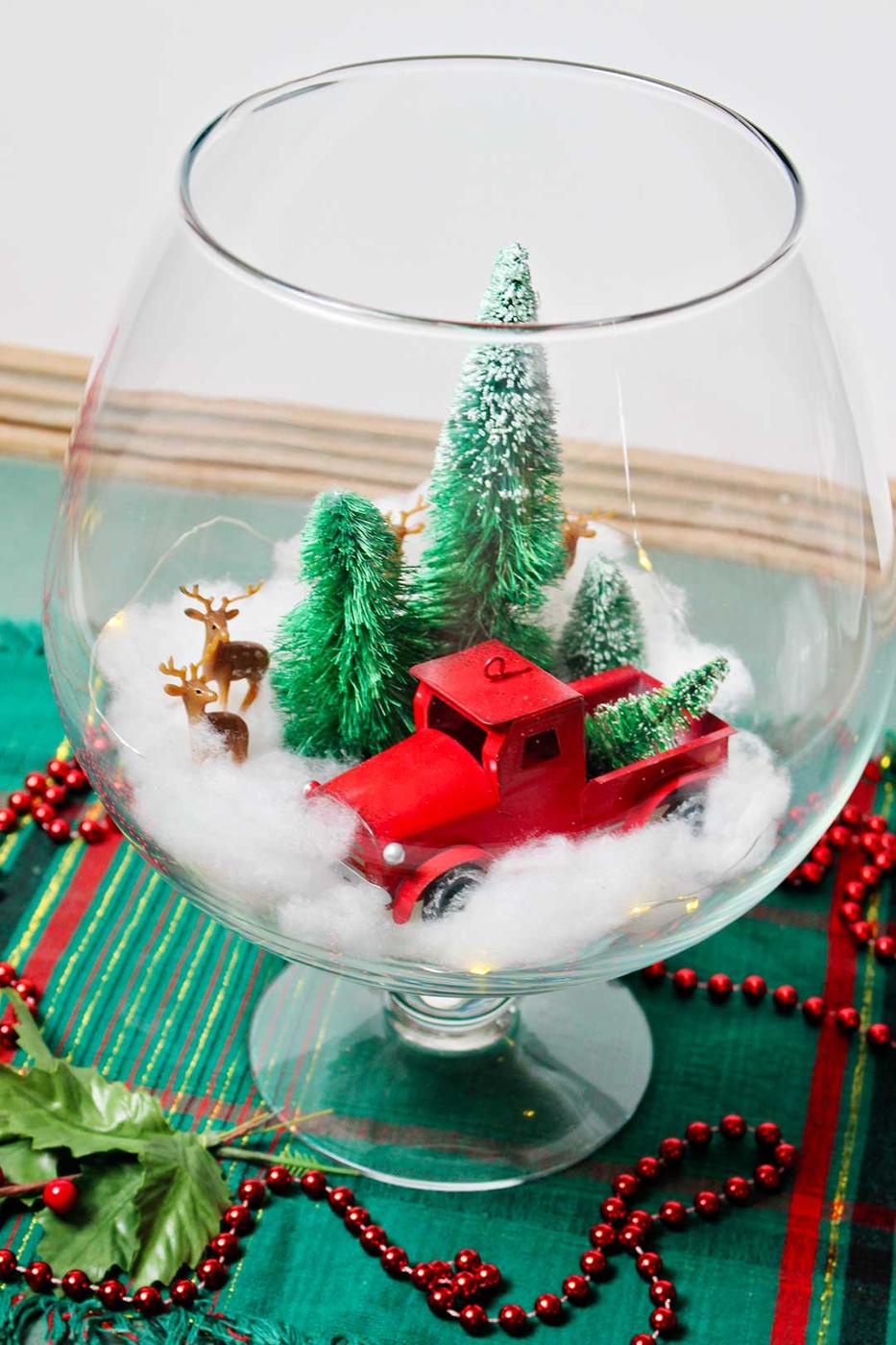 A tall glass bowl holding bottle brush trees, a red truck ornament, a tiny deer and fairy lights on a bed of white snow