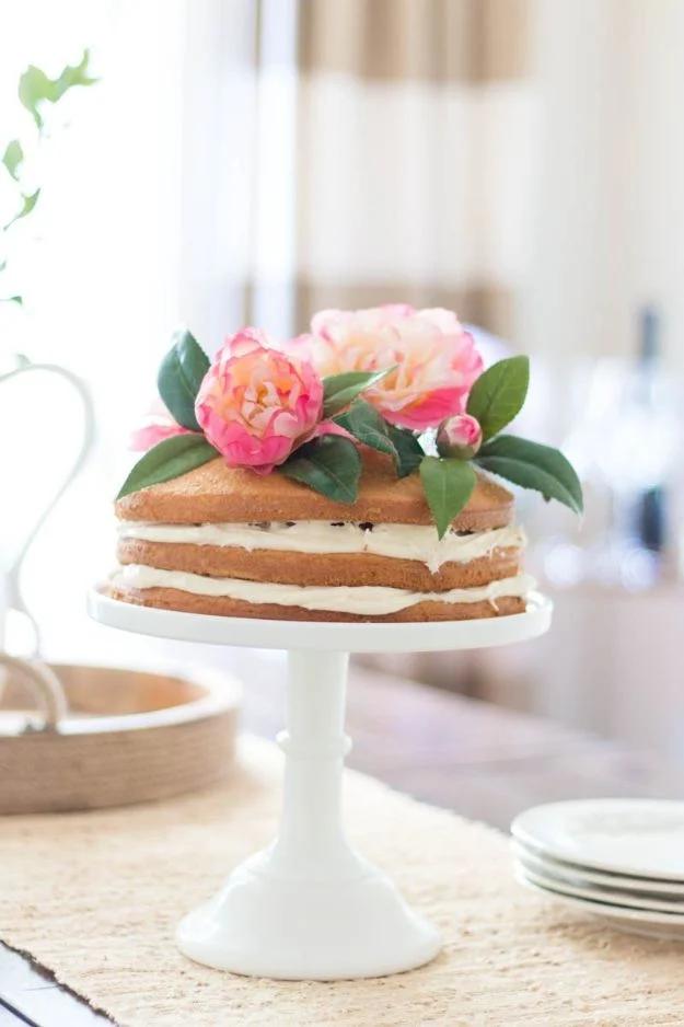 Naked layer cake on a white pedestal stand topped with pink faux peonies and green leaves, pale styled table behind