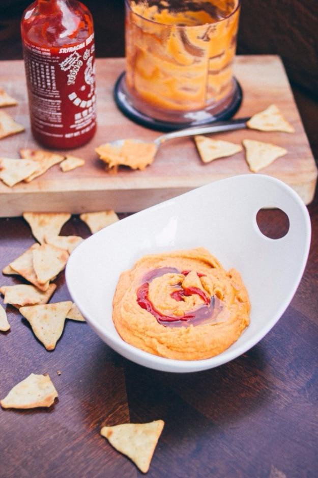 Creamy sriracha hummus party dip swirled in a bowl with olive oil, paprika and pita chips around the edge