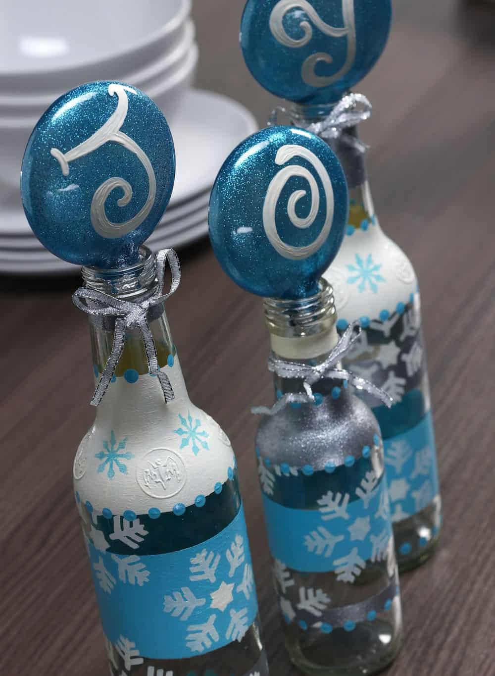 Three bottles painted in blue, silver and white stripes and dots with glitter ornaments spelling JOY hanging from their necks