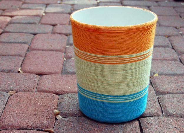 Trash can wrapped in bands of orange, cream and turquoise yarn, standing outdoors on red brick pavers