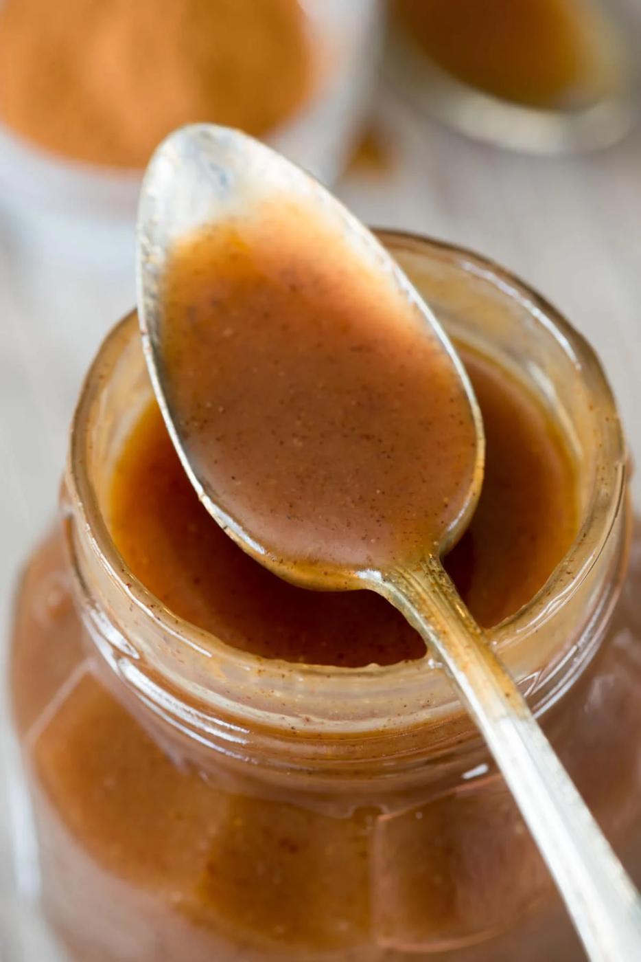 Glass jar of golden cinnamon caramel sauce with a spoon lifting a thick ribbon above the rim on white marble