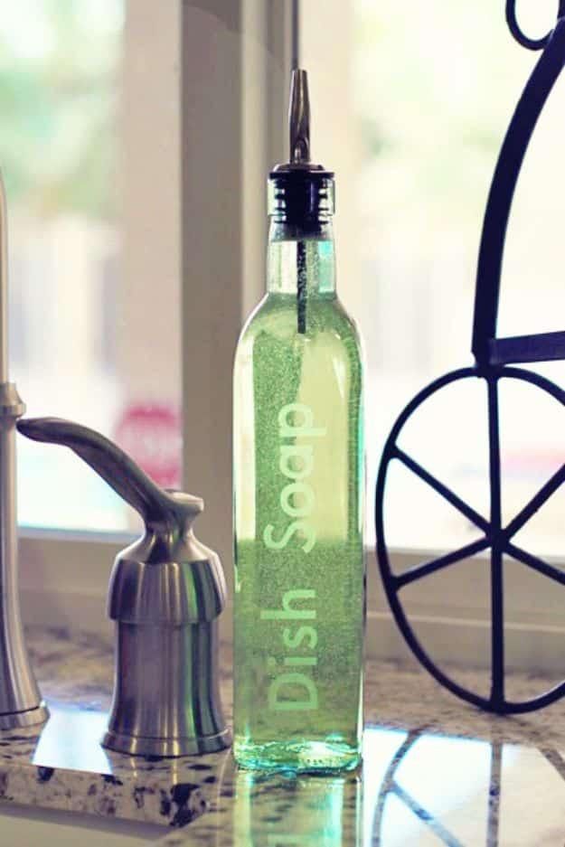 Tall glass olive oil bottle of green dish soap etched with the words dish soap and fitted with a metal pour spout